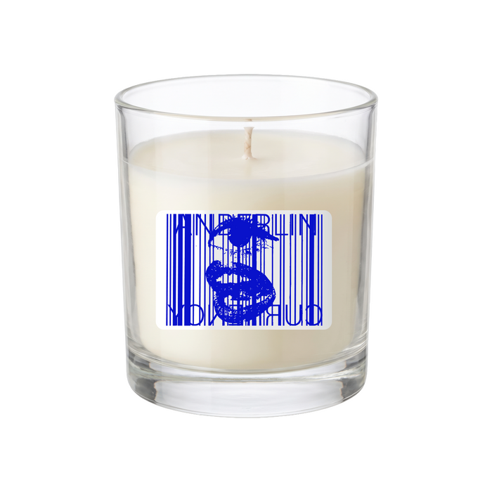 A white candle in a clear glass holder features a label with a blue barcode design and an abstract face, making it a collectible limited edition piece.