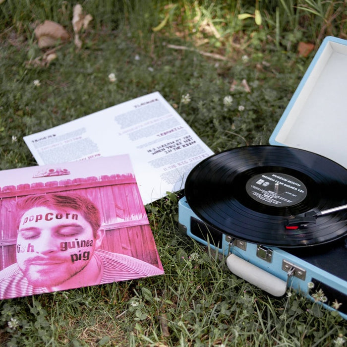 A portable blue turntable sits on grass, spinning a vinyl record. Next to it is the Museum Mouth album cover featuring a man's face with text, plus an open lyric sheet. White clover flowers are scattered nearby.