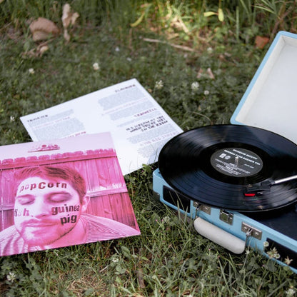 A portable blue turntable sits on grass, spinning a vinyl record. Next to it is the Museum Mouth album cover featuring a man's face with text, plus an open lyric sheet. White clover flowers are scattered nearby.