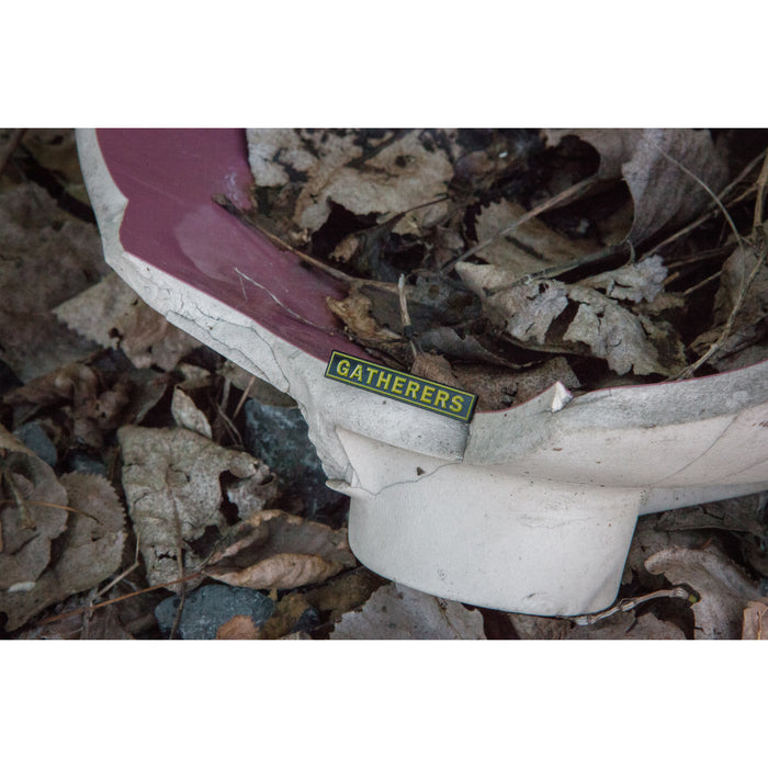 A broken white and red object lies among dry leaves, featuring a small yellow and black Gatherers label—reminiscent of an Alive Beyond Repair enamel pin by Gatherers left behind.